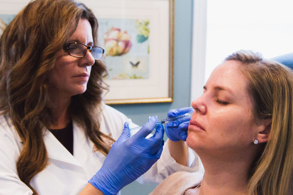 Provider wearing gloves carefully places dermal filler in Mason, OH, into patient's cheek. The patient leans back and remains calm with her eyes closed.