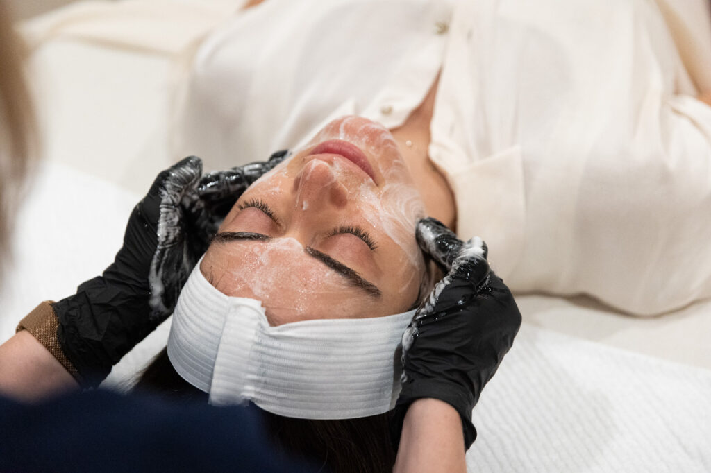 Patient lying on her back wearing a robe as the provider wearing gloves massages a facial cleanser into her cheeks. This is a treatment that pairs well with dermal fillers in Mason, OH, to help patients maintain a youthful, radiant glow.